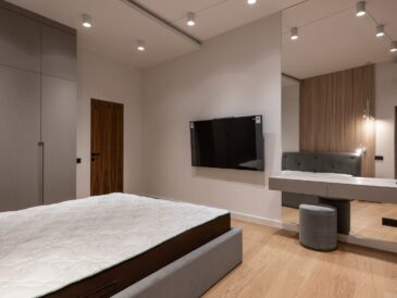 Comfortable bed and wardrobe in modern bedroom placed in front of TV hanging on wall near dressing table with big mirror