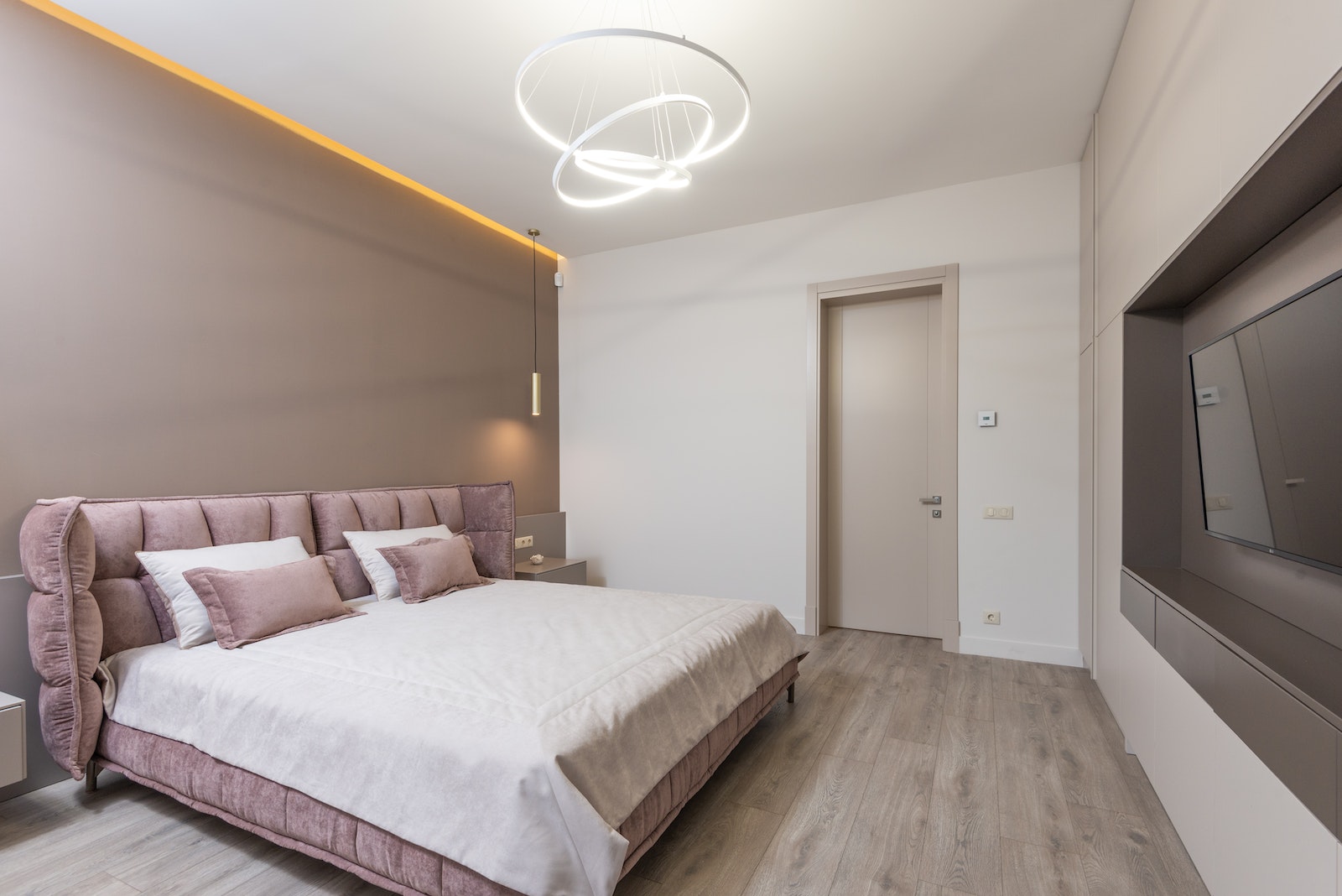Interior design of comfortable bedroom with light brown and white painted walls furnished with bed with pink upholstery decorated with pillows Illuminated by modern chandelier